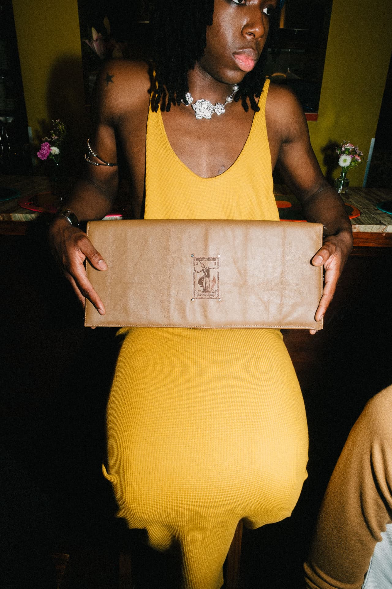 Model in a yellow ribbed dress holding a tan leather clutch stamped with a Khmer motif.
